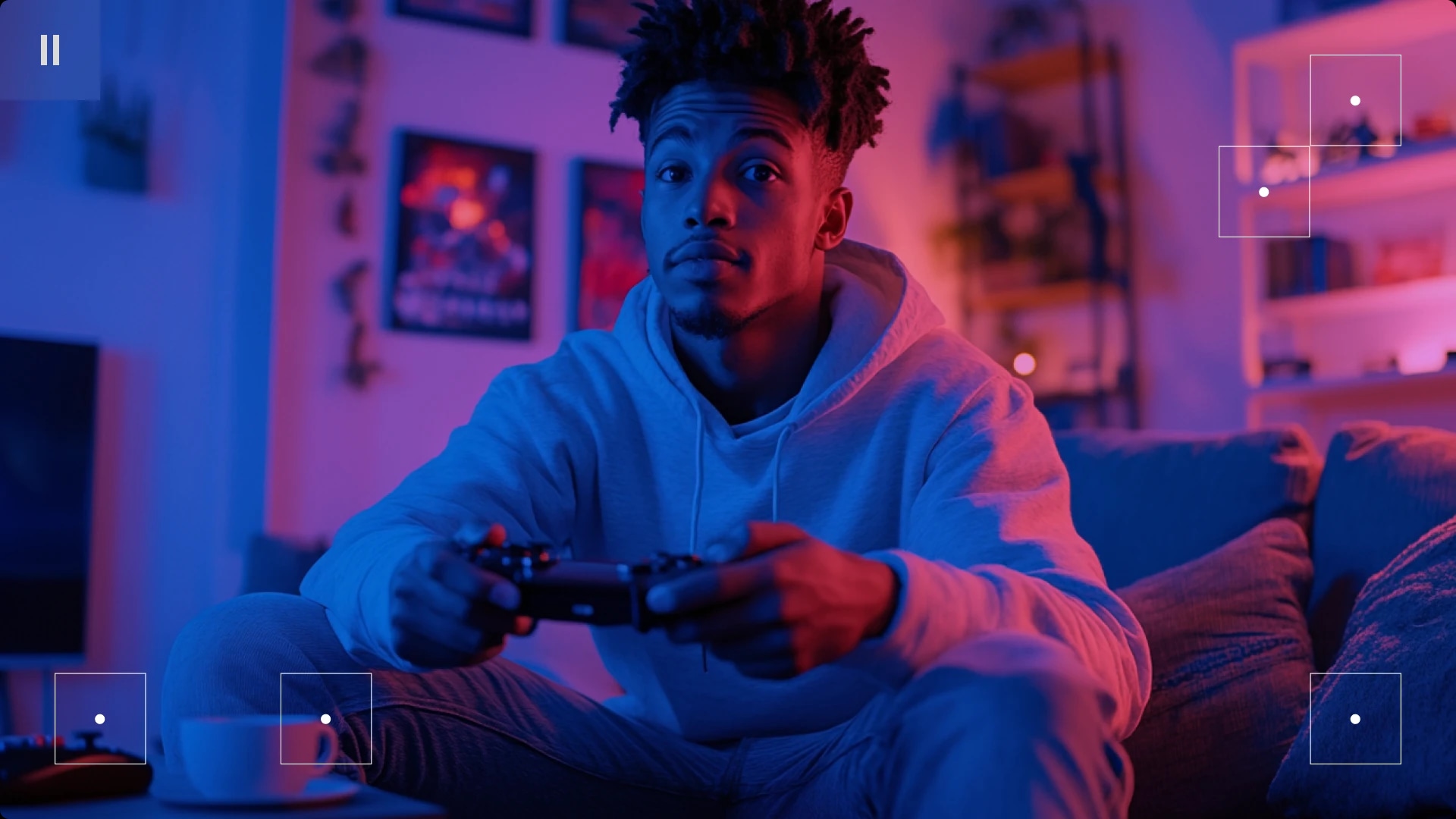 A young man sitting on a couch, holding a game controller, in a room illuminated with blue and pink lighting.