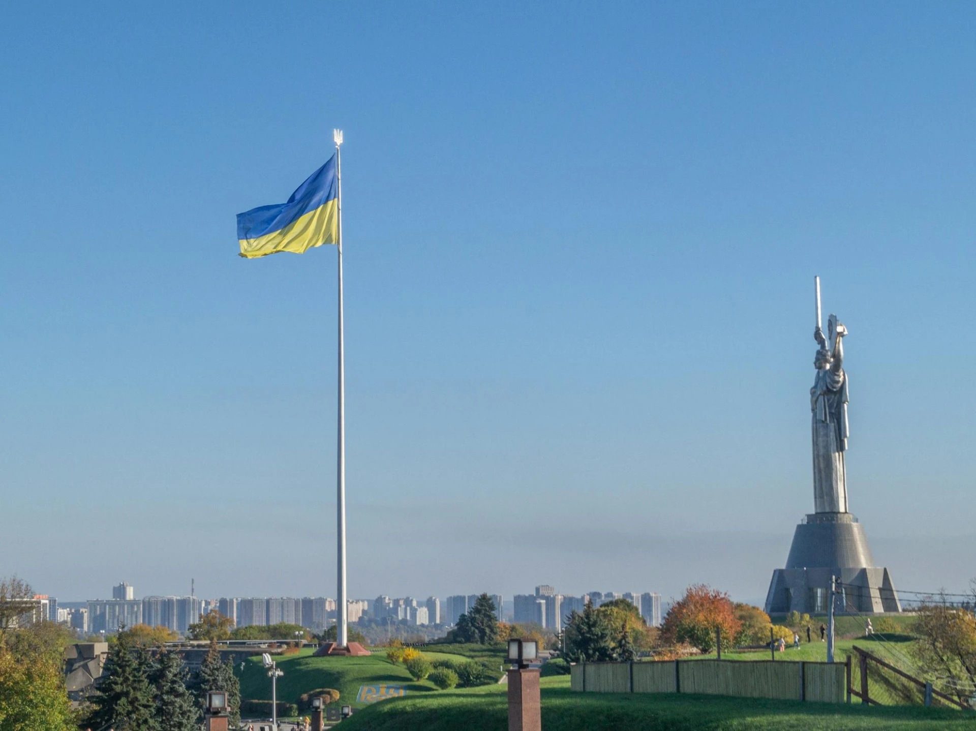 Ukrainian flag flying near the Statue of Liberty in a park with a city skyline in the background.