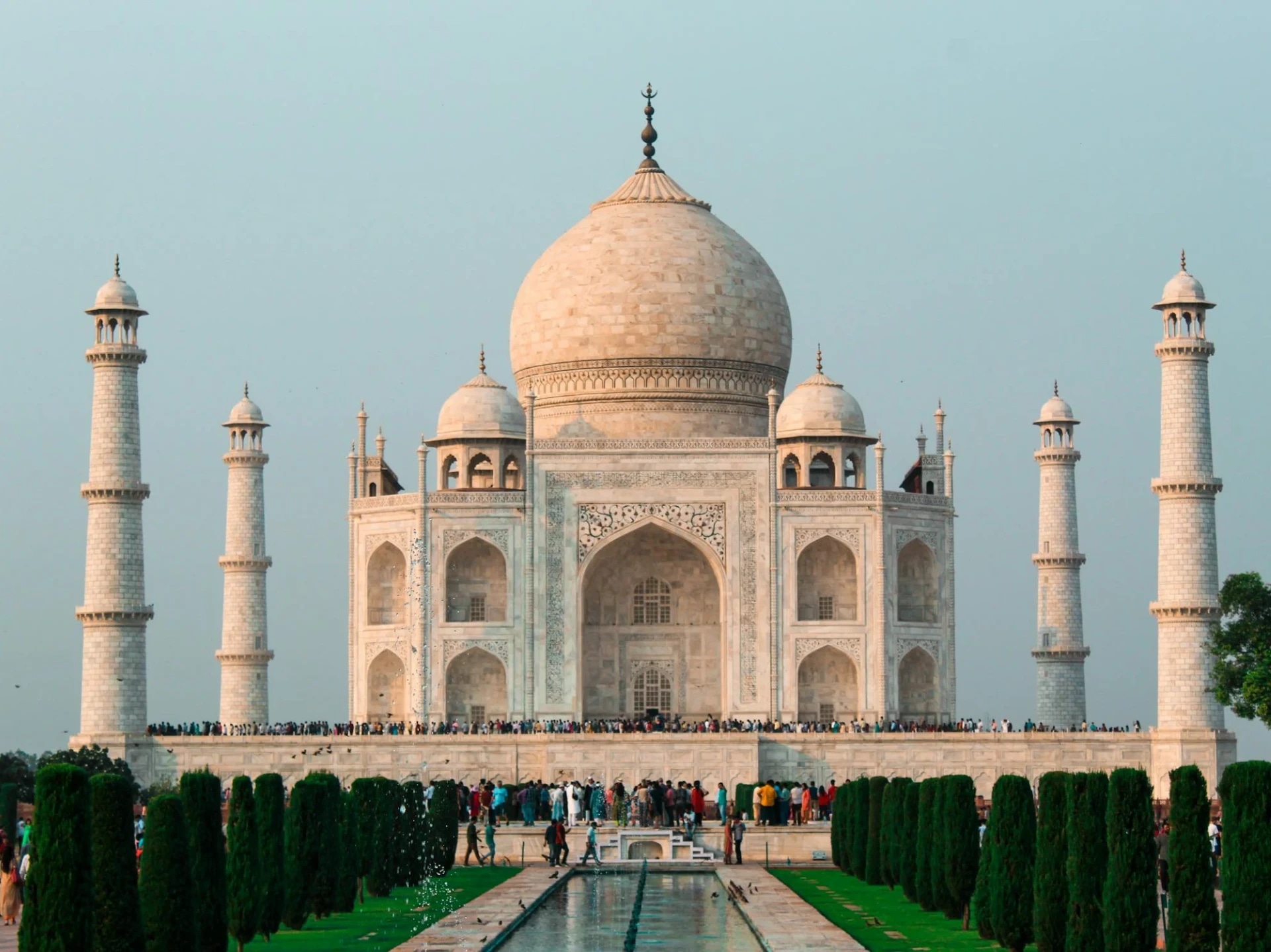 The Taj Mahal with a large crowd of visitors in front, surrounded by green gardens and a reflecting pool.