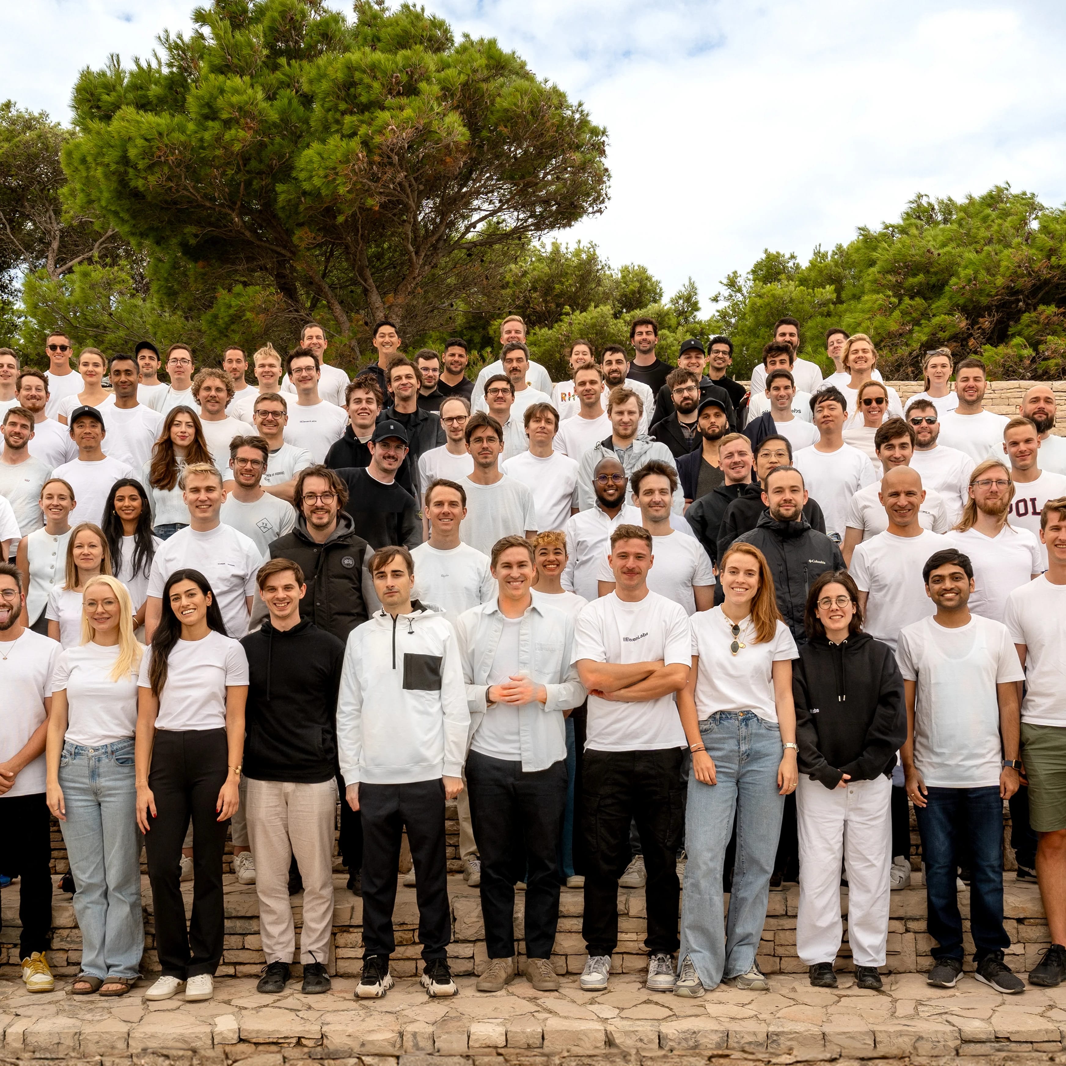 Group of people standing outdoors on steps, smiling, with trees and cloudy sky in the background.