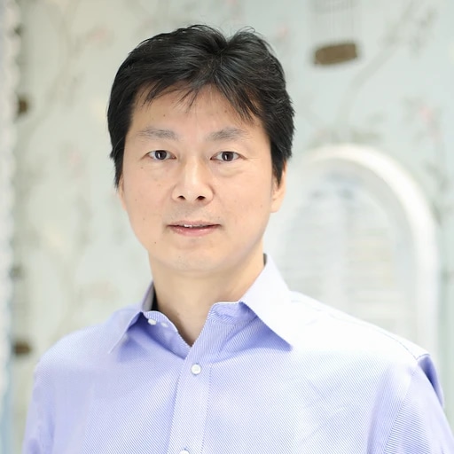 A man with short black hair wearing a light blue dress shirt, standing indoors with a blurred background.