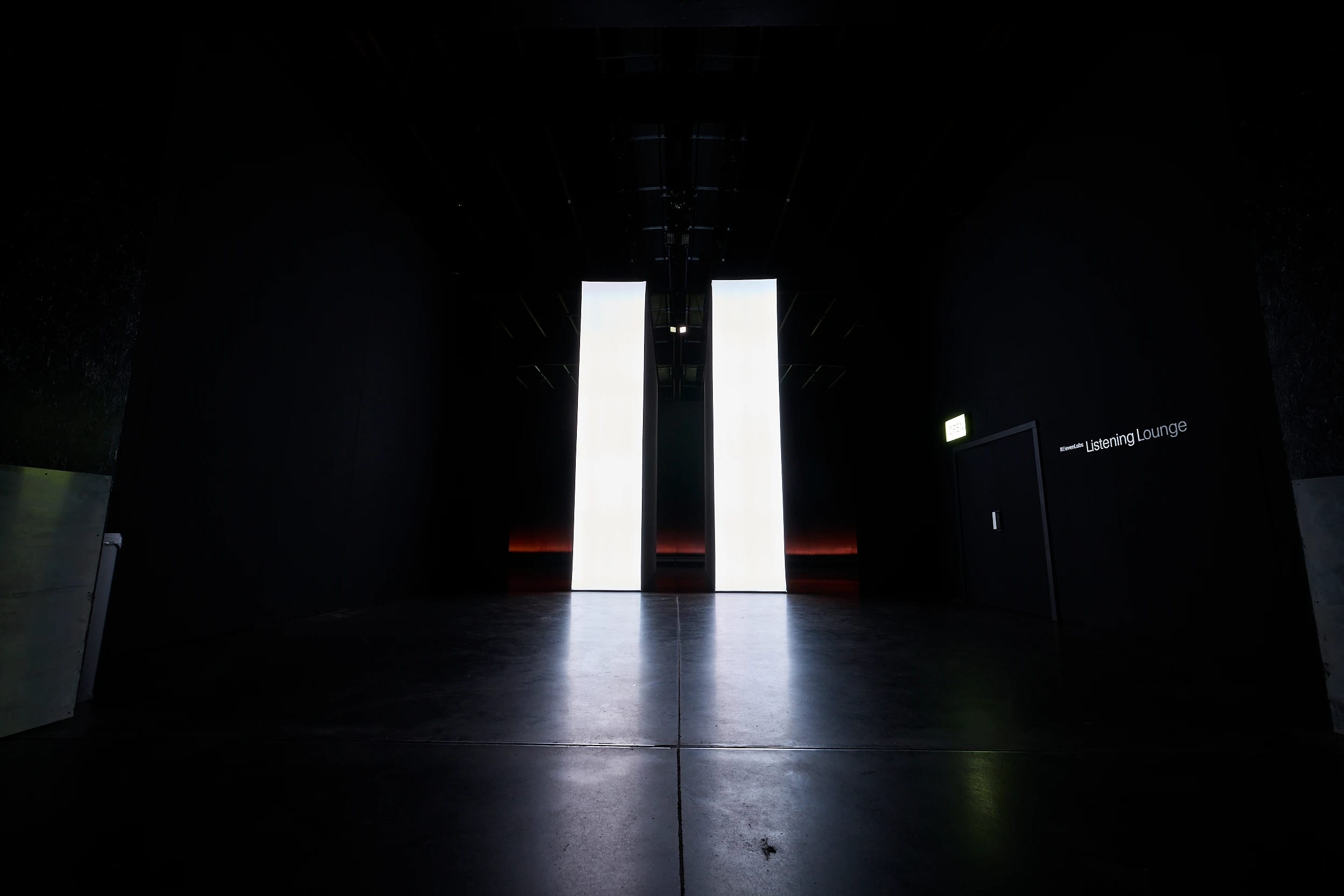 Dark exhibition space with two glowing panels beside the entrance to the Listening Lounge.