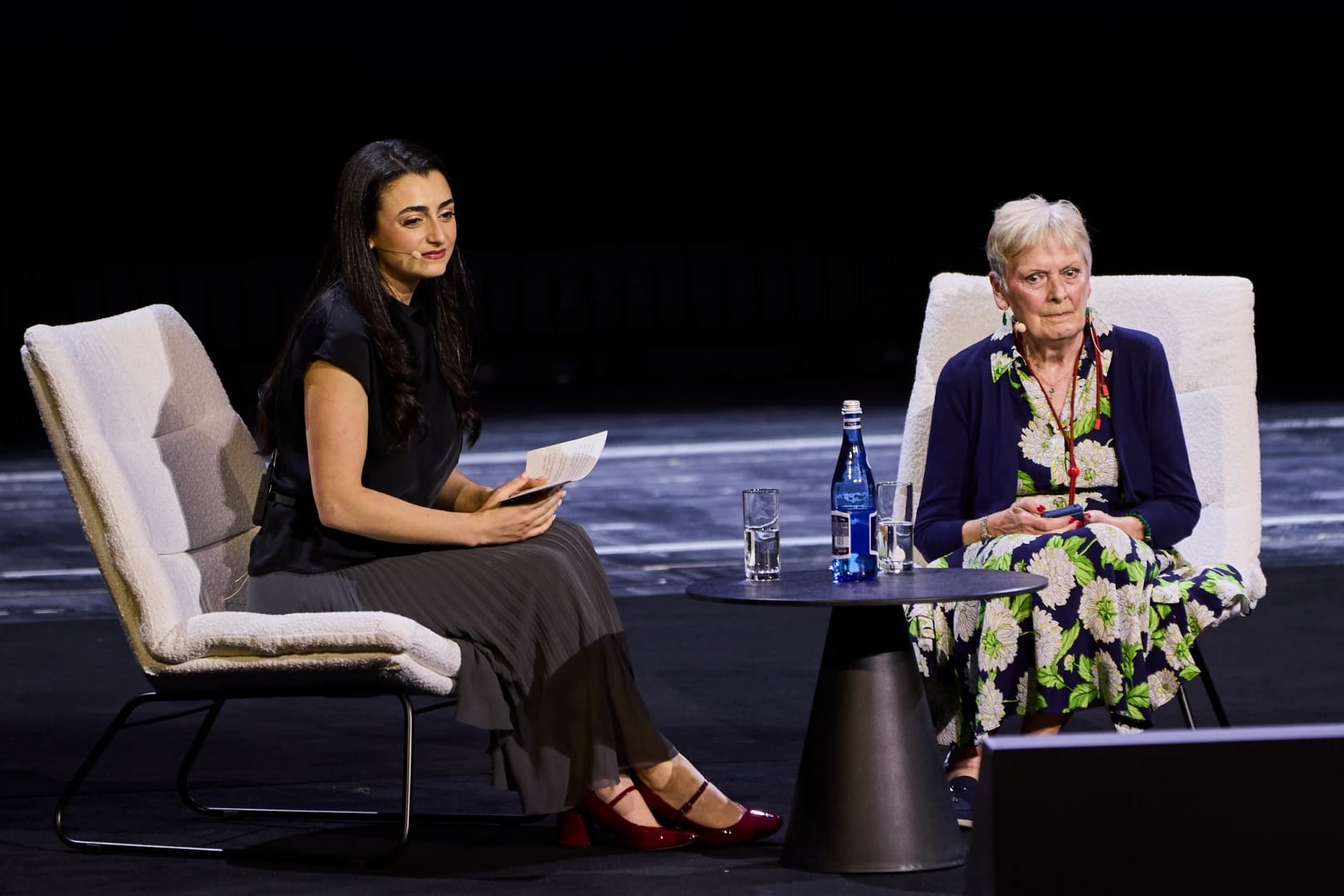 Two women seated on stage during a discussion, one holding a paper, glasses and water on table.