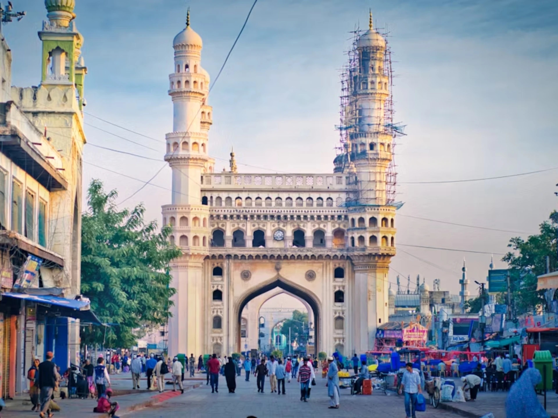 A busy street scene in front of a large, ornate gateway with two minarets, some under construction scaffolding, and people walking and shopping.