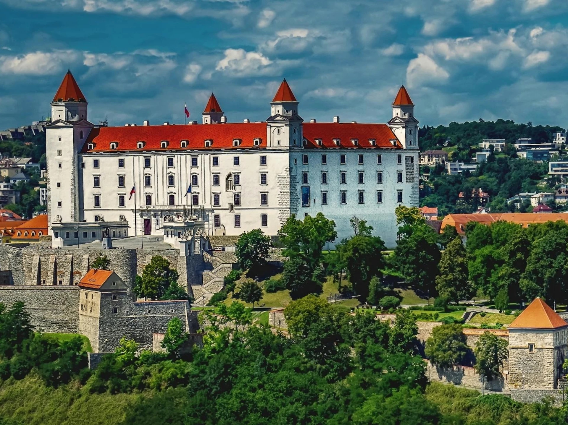 A large white castle with red roofs and four towers, surrounded by green trees and a stone wall, under a partly cloudy sky.