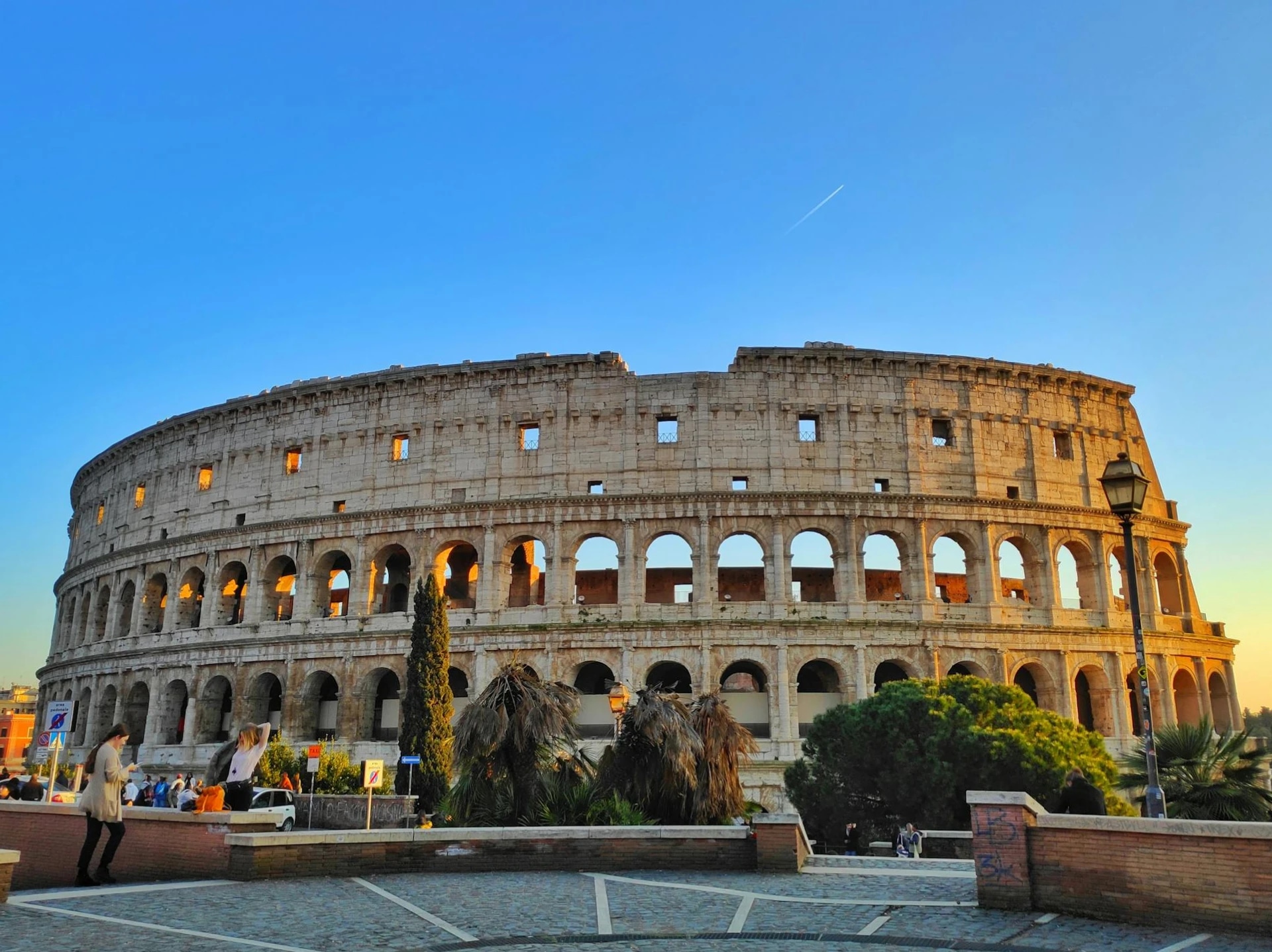The Colosseum in Rome during sunset with people walking nearby and a clear blue sky.