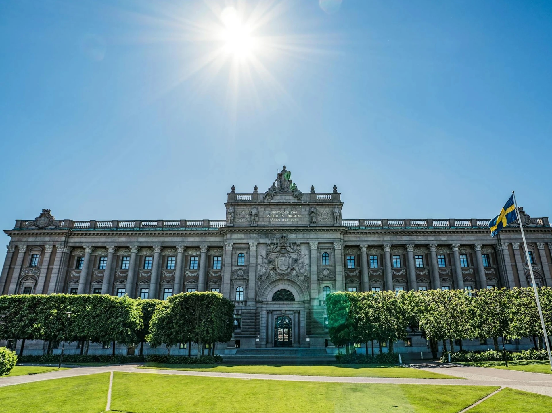 A grand historic building with a Swedish flag, surrounded by green trees and a well-maintained lawn under a bright, sunny sky.