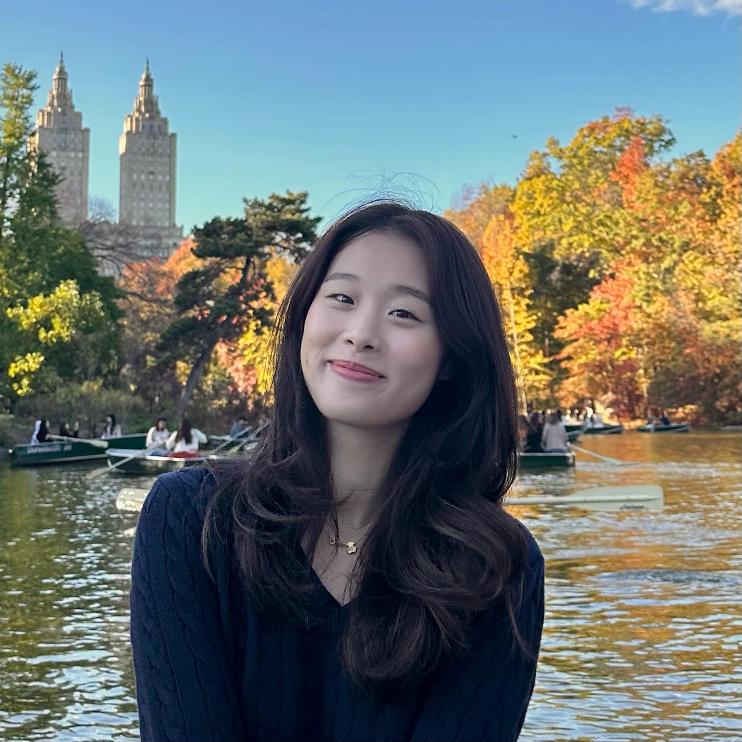 A smiling woman with long hair at a lake surrounded by autumn trees and boats.