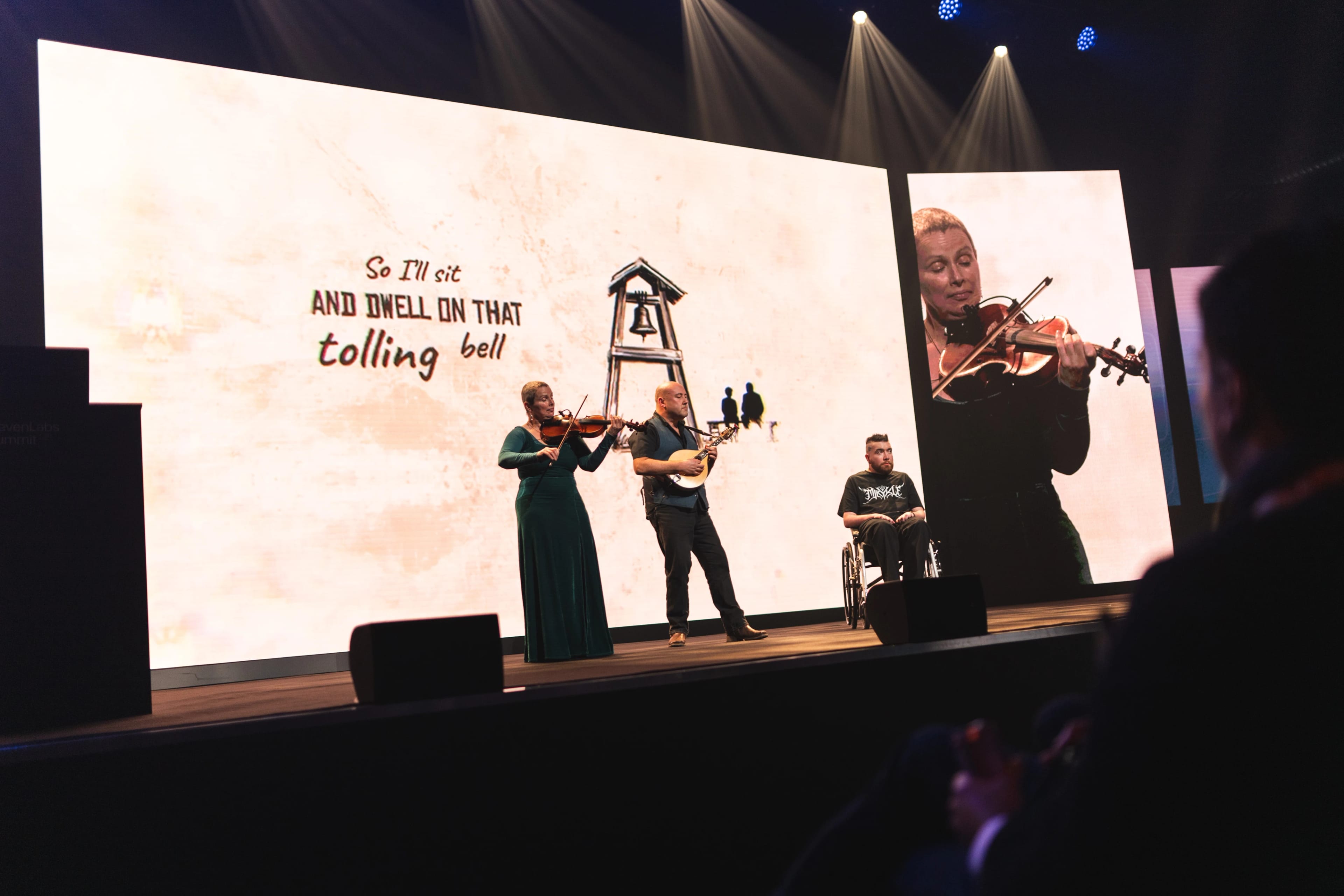A man in a wheelchair sits onstage while a violinist and mandolinist play, behind him is a screen that says "So I sit and dwell on that tolling bell"
