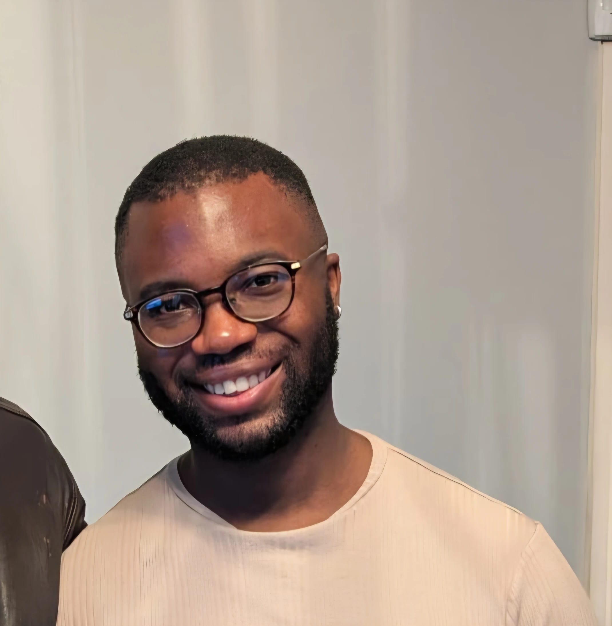 A person smiling, wearing glasses and a light-colored shirt, standing against a neutral background.
