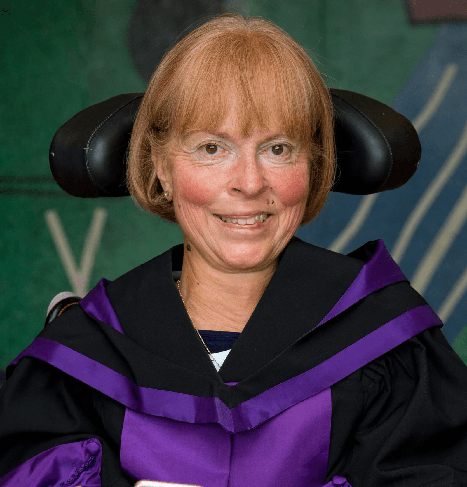 Smiling Baroness Campbell of Surbiton in a wheelchair wearing black and purple academic robes.